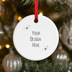 Puede incluir: Un adorno navideño blanco y redondo con el texto "YOUR DESIGN HERE" en gris. El adorno tiene diseños de estrellas y una cinta roja para colgar. El fondo está borroso con decoraciones navideñas.