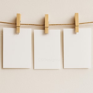 Peut inclure: Trois cartons blancs vierges sont accrochés à une ficelle marron avec des pinces à linge en bois. Les cartons sont rectangulaires et suspendus sur un fond de couleur neutre. Les pinces à linge sont de couleur bois clair.