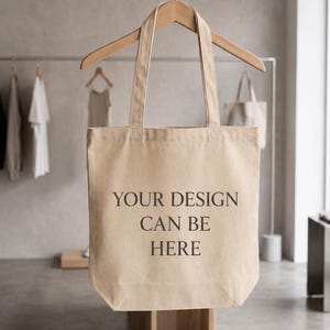 May include: A beige canvas tote bag hanging on a wooden hanger. The bag features two long shoulder straps and the black printed text "YOUR DESIGN CAN BE HERE" centered on the front. The material has a visible woven texture and a rectangular shape.