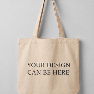 May include: A beige canvas tote bag with two long shoulder straps. The front of the bag features the black printed text "YOUR DESIGN CAN BE HERE" centered in a simple serif font. The bag is constructed from a plain, light-colored fabric material.