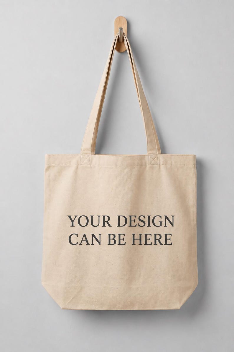 May include: A beige canvas tote bag with two long shoulder straps. The front of the bag features the black printed text "YOUR DESIGN CAN BE HERE" centered in a simple serif font. The bag is constructed from a plain, light-colored fabric material.