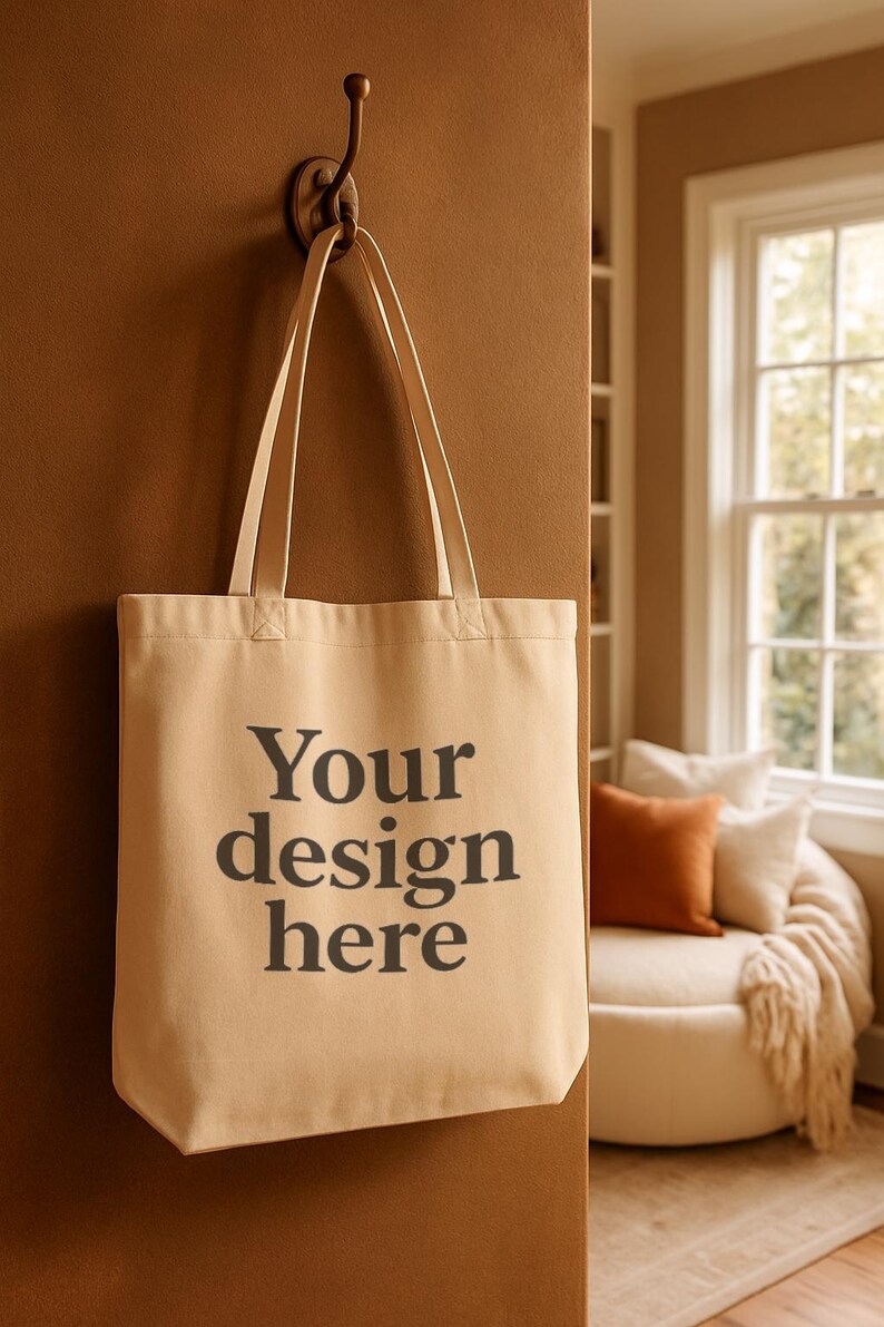 Może przedstawiać: Beżowa torba z nadrukiem "Your design here" w kolorze czarnym. Torba wisi na brązowym haczyku na brązowej ścianie. Tło obejmuje okno i część wypoczynkową.