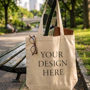 May include: A beige canvas tote bag with two shoulder straps featuring the black text YOUR DESIGN HERE printed on the front. A pair of tortoiseshell eyeglasses is hooked onto the left strap, and a brown leather notebook sits inside the bag.