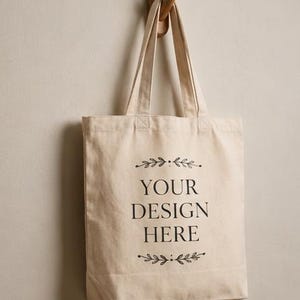 May include: A beige canvas tote bag with two shoulder straps, featuring the black printed text "YOUR DESIGN HERE" centered on the front, framed by decorative black leaf illustrations above and below the text, hanging from a wooden wall hook.