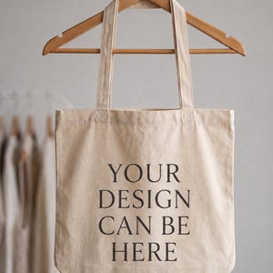 May include: A beige canvas tote bag hanging from a wooden hanger. The bag features the black printed text "YOUR DESIGN CAN BE HERE" centered on the front. The tote has two long fabric handles and a rectangular shape with a flat bottom edge.