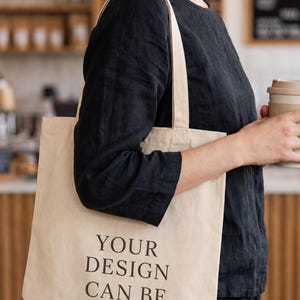 May include: A beige canvas tote bag with two shoulder straps, featuring the black printed text "YOUR DESIGN CAN BE HERE" on the front. The bag is constructed from a light-colored fabric and is designed to be carried over the shoulder.