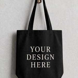 May include: A black tote bag with two shoulder straps, featuring the text "YOUR DESIGN HERE" printed in a light cream color in the center. The bag is constructed from a solid fabric material and is displayed hanging from a wooden wall hook.