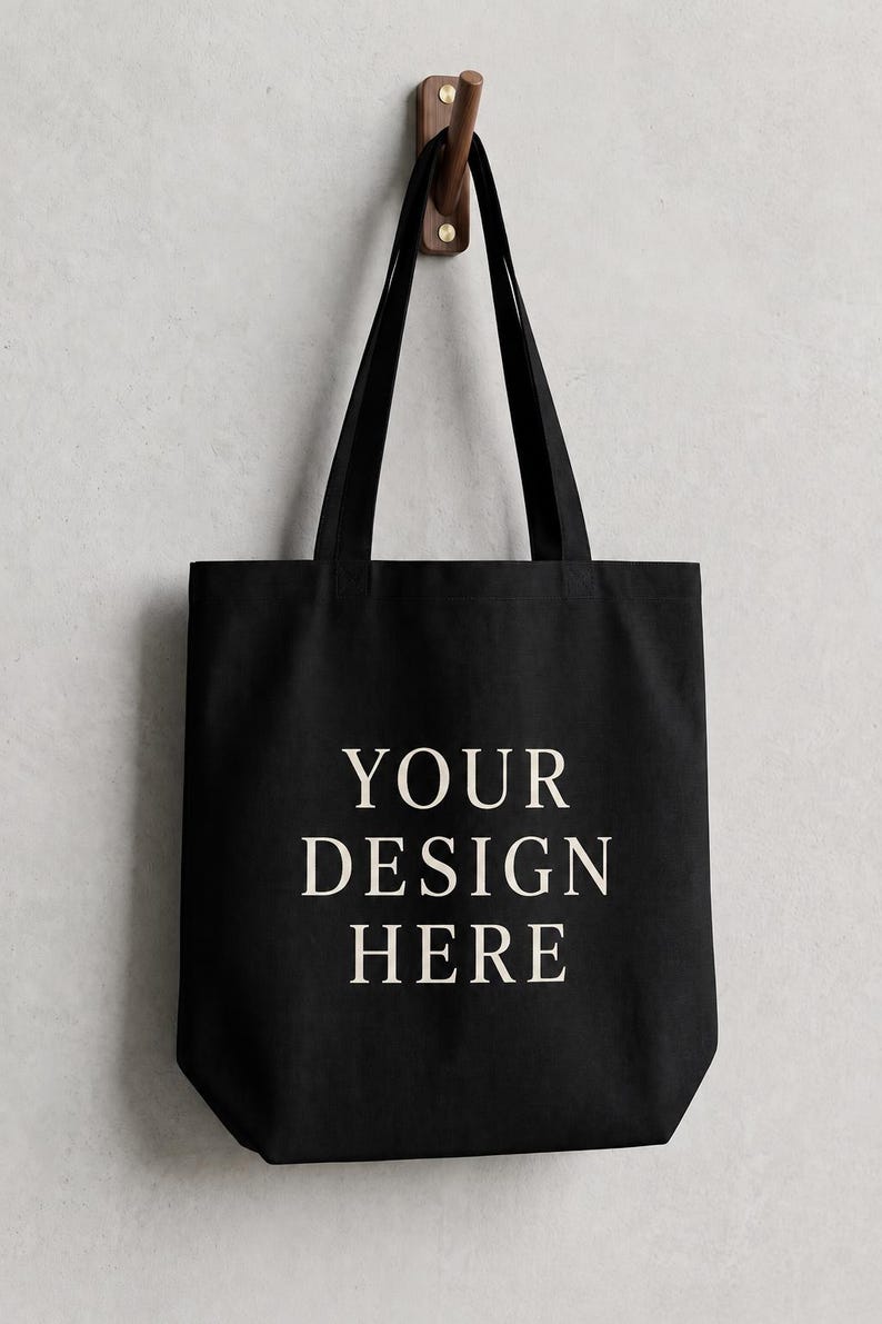May include: A black tote bag with two shoulder straps, featuring the text "YOUR DESIGN HERE" printed in a light cream color in the center. The bag is constructed from a solid fabric material and is displayed hanging from a wooden wall hook.