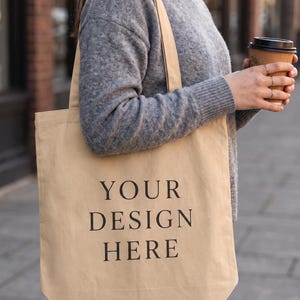 May include: A beige canvas tote bag featuring the black text "YOUR DESIGN HERE" printed in a centered, serif font. The bag has two shoulder straps and a rectangular shape, providing a neutral surface for custom graphics or branding.