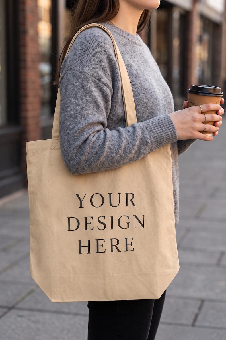 May include: A beige canvas tote bag featuring the black text "YOUR DESIGN HERE" printed in a centered, serif font. The bag has two shoulder straps and a rectangular shape, providing a neutral surface for custom graphics or branding.