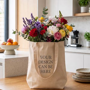 May include: A beige canvas tote bag with two handles and the text "YOUR DESIGN CAN BE HERE" printed in black on the front. The bag is filled with a large, colorful bouquet of mixed flowers including red roses, white peonies, and purple lavender.