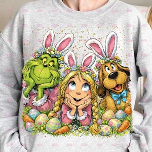 May include: A light gray sweatshirt featuring a Grinch character, a girl, and a dog, all wearing bunny ears and flower crowns. Colorful Easter eggs and carrots are scattered around the characters, creating a festive holiday design.