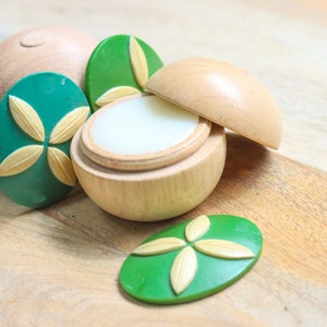 May include: A wooden container with a lid, containing a white balm. The container is decorated with green and yellow floral designs.
