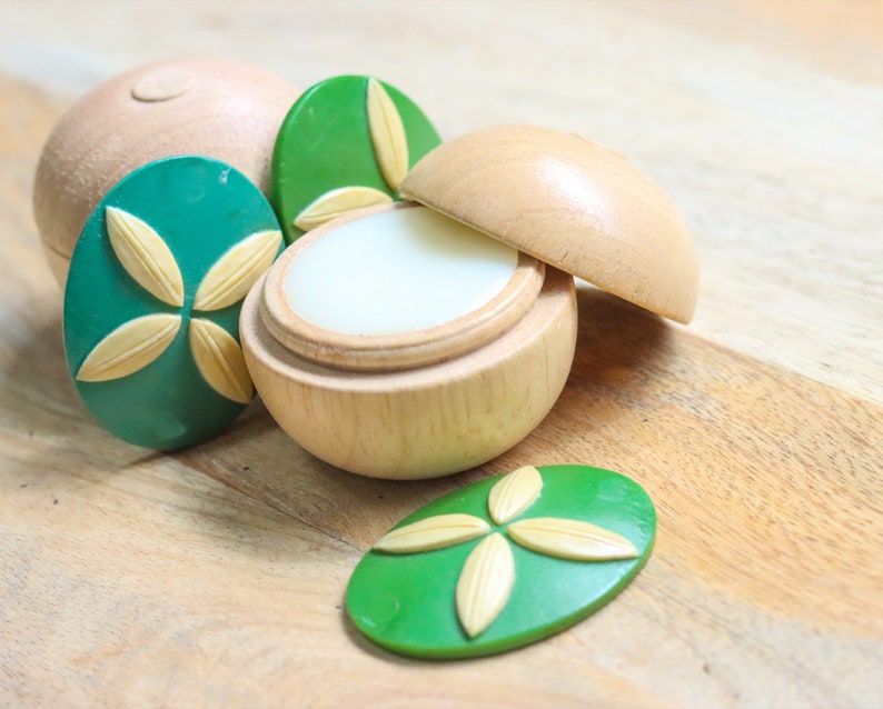 May include: A wooden container with a lid, containing a white balm. The container is decorated with green and yellow floral designs.