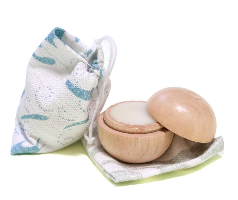 May include: A wooden container with a lid, filled with a white balm, is sitting on a light green and white fabric. The container is next to a small, white drawstring bag with a blue and white pattern.