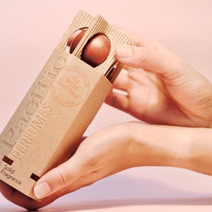 May include: A brown cardboard box with the text "Pacific Perfumes" and "The Wild Goddess" printed on it. The box contains solid fragrance in the form of small, round, brown discs. The box is being held by a person's hands.