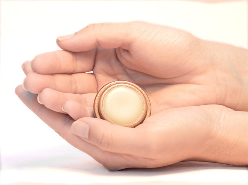 May include: A small round container with a wooden lid, containing a white substance, held in two hands.
