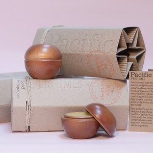 May include: Two wooden containers of solid fragrance, one open, with a light brown lid and a yellow substance inside. The containers are on a brown cardboard box with the text "Pacific Perfumes" and "The Wild Goddess" printed on it. There is a smaller brown cardboard box with the text "Solid Fragrance" printed on it. There is a white card with the text "Pacific Perfumes" and a description of the product.