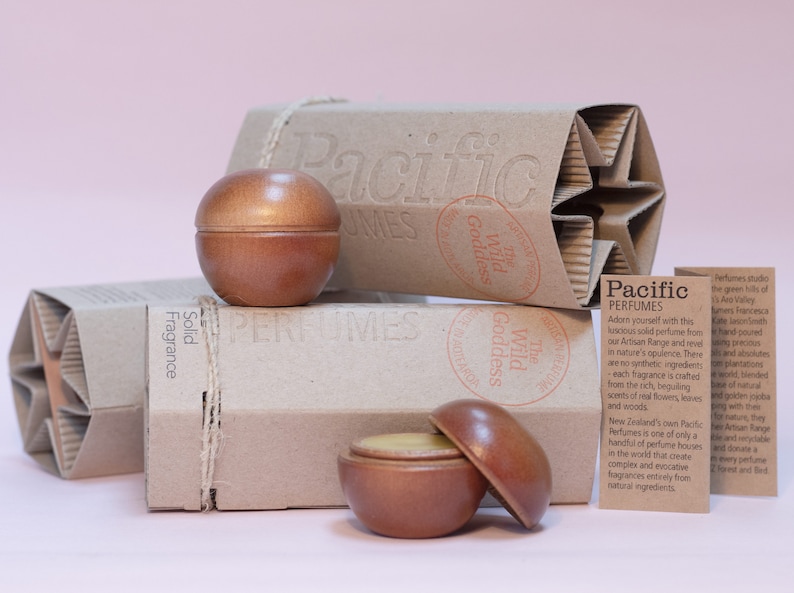 May include: Two wooden containers of solid fragrance, one open, with a light brown lid and a yellow substance inside. The containers are on a brown cardboard box with the text "Pacific Perfumes" and "The Wild Goddess" printed on it. There is a smaller brown cardboard box with the text "Solid Fragrance" printed on it. There is a white card with the text "Pacific Perfumes" and a description of the product.