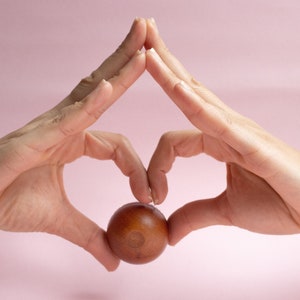 May include: Two hands forming a heart shape with a brown wooden ball in the center, against a pink background.