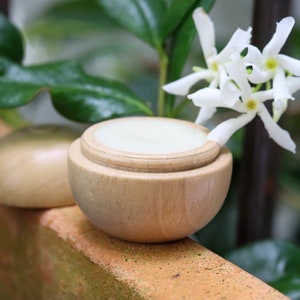 May include: A wooden container with a lid, filled with a white cream, sits on a brown surface. A white jasmine flower with green leaves is in the background.