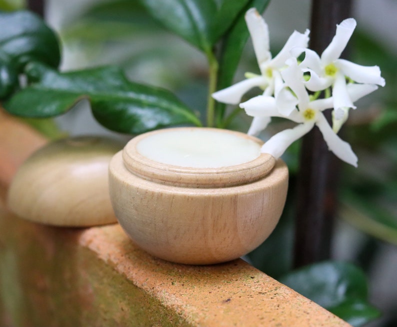 May include: A wooden container with a lid, filled with a white cream, sits on a brown surface. A white jasmine flower with green leaves is in the background.