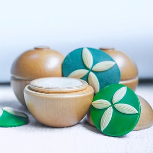 May include: Three wooden containers with lids, two are round and one is a smaller square shape. The round containers have green lids with a white flower design. The square container has a white lid.