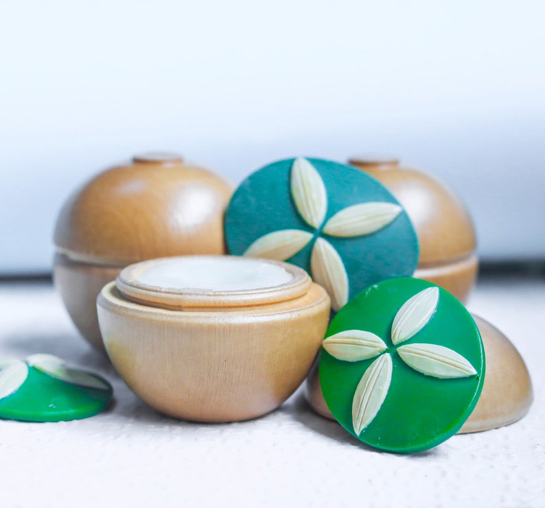 May include: Three wooden containers with lids, two are round and one is a smaller square shape. The round containers have green lids with a white flower design. The square container has a white lid.