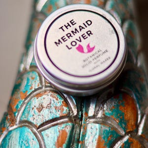 Könnte beinhalten: Kleine, runde Dose mit weißem Deckel und dem Text "THE MERMAID LOVER" in Schwarz sowie einer rosa Meerjungfrauen-Schwanz-Grafik. Die Dose liegt auf einer strukturierten, türkis- und kupferfarbenen Oberfläche.