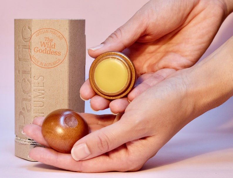 May include: A wooden box with the text "The Wild Goddess" and "Artisan Perfume" printed on it. The box is next to a small wooden container with a yellow balm inside. A wooden ball is also shown.