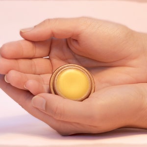May include: A small round container of yellow balm in a wooden container with a gold rim.