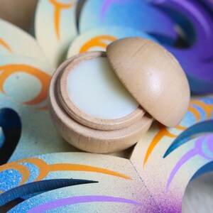 May include: A small wooden container with a lid, open to reveal a white balm inside. The container is sitting on a colorful, abstract background.