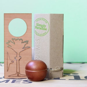 May include: A brown wooden box with a white label that says "Artisan Perfume Simply Paradise Made in Aotearoa". The box is sitting on a brown surface next to a brown wooden round container and a wooden cutout of a tree.