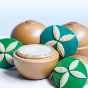 May include: A set of four wooden containers with green lids featuring a white flower design. The containers are round and have a smooth, polished finish. One container is open, revealing a white, creamy substance inside.