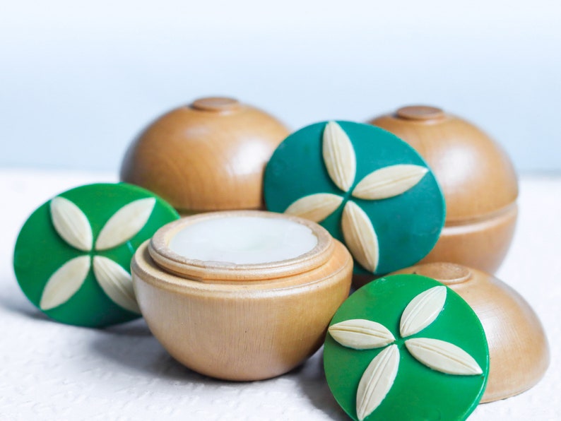 May include: A set of four wooden containers with green lids featuring a white flower design. The containers are round and have a smooth, polished finish. One container is open, revealing a white, creamy substance inside.