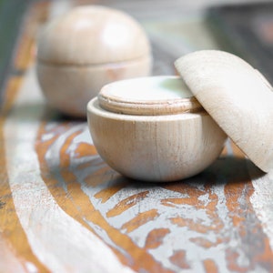 May include: Two wooden containers with lids, one open revealing a white cream inside. The containers are on a rustic wooden surface with a light gray and brown paint design.