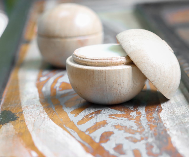 May include: Two wooden containers with lids, one open revealing a white cream inside. The containers are on a rustic wooden surface with a light gray and brown paint design.