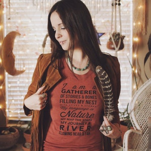 May include: A woman wearing a red t-shirt with a black graphic that reads "I am a gatherer of stories & bones filling my nest with sticks & stones. My language is Nature. My map the stars. My journey a river flowing near & far." She is holding a feather in her right hand.