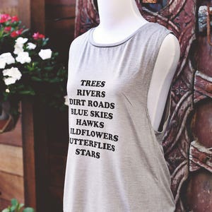Light grey muscle tank.  Inspired by our love of Texas.  Says, Trees Rivers Dirt Roads Blue Skies Hawks Wildflowers Butterflies Stars on front int black ink.