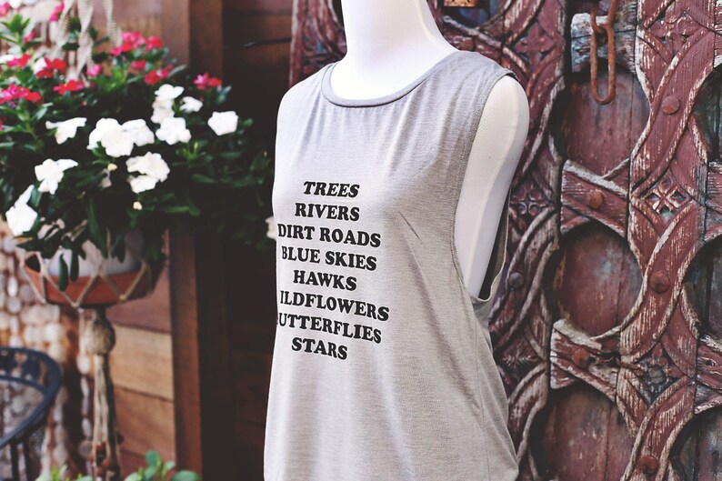 Light grey muscle tank.  Inspired by our love of Texas.  Says, Trees Rivers Dirt Roads Blue Skies Hawks Wildflowers Butterflies Stars on front int black ink.