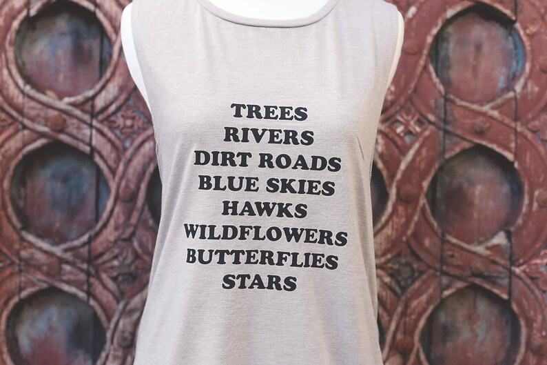 Light grey muscle tank.  Inspired by our love of Texas.  Says, Trees Rivers Dirt Roads Blue Skies Hawks Wildflowers Butterflies Stars on front int black ink.