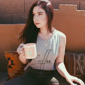 May include: A woman wearing a gray tank top with the word "Coffee" written on it, holding a pink and blue ceramic mug. She is sitting on a colorful patterned cushion.