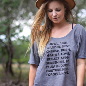 May include: A woman wearing a gray t-shirt with the text "HOWL. SEEK. WANDER. NEST. CHERISH. BURN. GATHER. LOVE. REFLECT. SING. SURRENDER. BE. RELEASE. GUIDE. NURTURE. HELP. FORGIVE. HOPE." printed on it.
