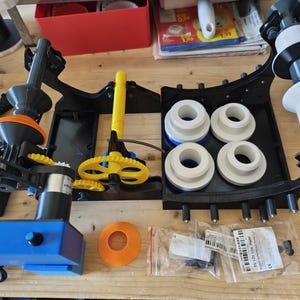 Può includere: Una macchina etichettatrice meccanica con base blu, telaio nero, ingranaggi gialli e rulli bianchi. Il dispositivo include un cavo di alimentazione, un componente circolare arancione e due bustine trasparenti contenenti minuteria ed un'etichetta.