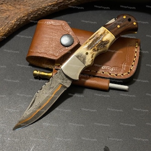 May include: A handcrafted hunting knife with a textured blade and a handle made of wood and antler. The knife is displayed with a brown leather sheath and a small sharpening rod. The blade has a unique, rustic appearance.