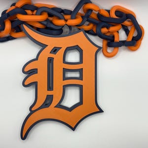 May include: An orange and navy blue Detroit Tigers logo with a chain. The logo is a stylized letter "D" with a navy blue outline. The chain is composed of orange and navy blue links.