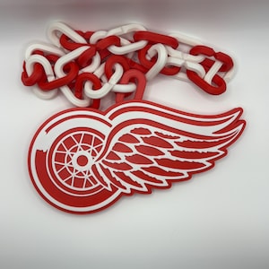 Op de afbeelding: Een rood en wit logo met een gevleugeld wielontwerp, verbonden met een ketting van rode en witte schakels. Het logo is een gestileerd embleem, waarschijnlijk voor een sportteam of organisatie.