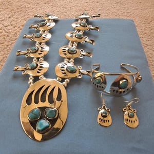 Könnte beinhalten: Ein silberfarbenes Schmuckset mit türkisfarbenen Steinen. Das Set umfasst eine Halskette mit einem Bärenklauen-Anhänger, ein Armreif und Ohrringe. Der Schmuck wird auf einer blauen Oberfläche präsentiert.