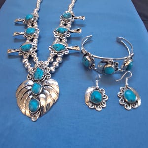 Puede incluir: Un conjunto de joyería de plata que incluye un collar tipo squash blossom, un brazalete y dos pendientes. Cada pieza presenta metal plateado pulido y piedras turquesas ovaladas incrustadas en un tono azul brillante.