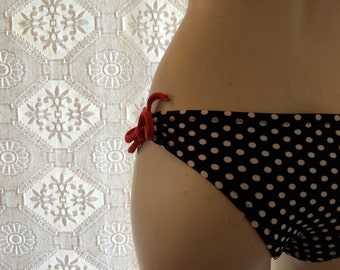 Puede incluir: Braguita de bikini negra con lunares blancos y lazos rojos laterales. El fondo es una tela blanca estampada.